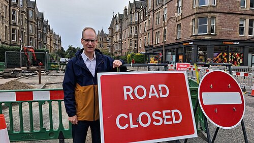 Councillor Neil Ross standing next to a "Road Closed" sign with buildings in the background